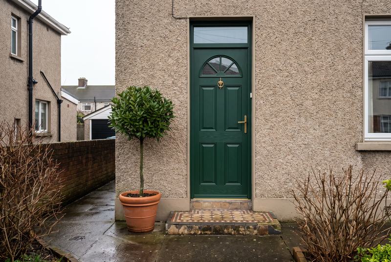 A dark green front door with a fanlight and brass handle on a pebble-dashed house, with a potted bay tree by the step