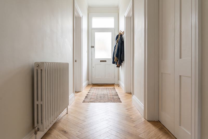 A long hall with an oak herringbone floor, a white radiator, white panelled doors and a glazed front door at the end