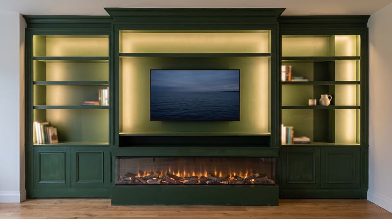 A green painted media wall with a wall-mounted TV in the centre recess, warm LED strips under the shelves, panelled cupboards and a long inset electric fire